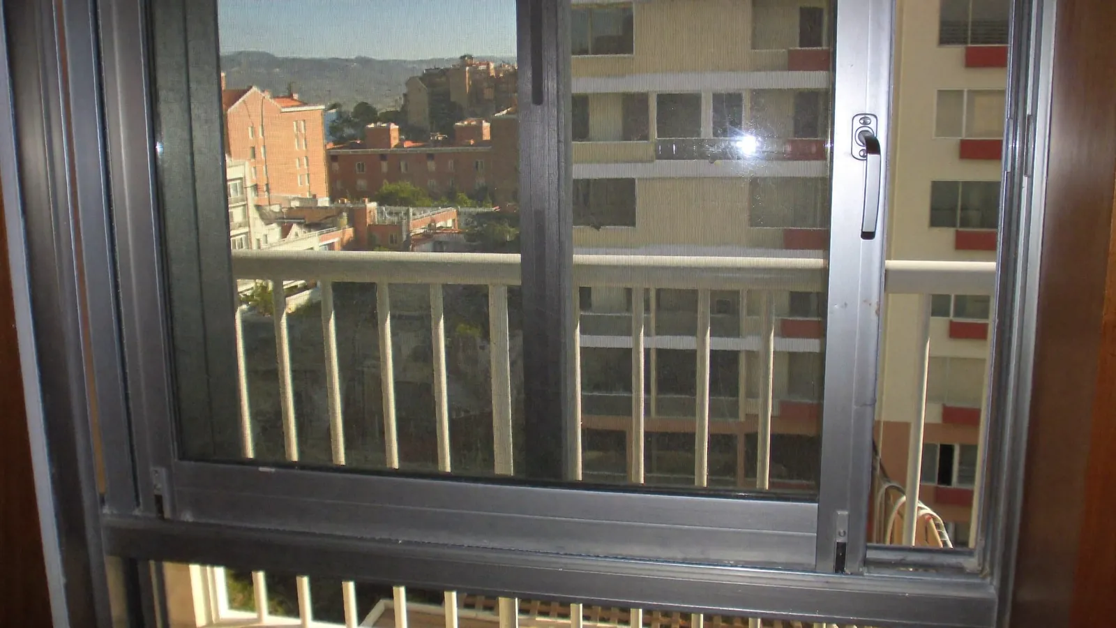 Corredera antigua de aluminio con vidrio sencillo que da a un balcón, con edificios de viviendas al fondo